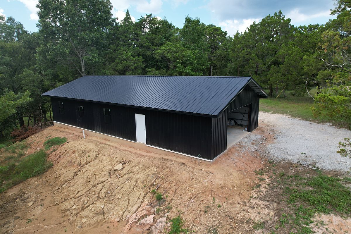 Black steel barndominium building on a cleared pad surrounded by woods in Coldspring, Texas
