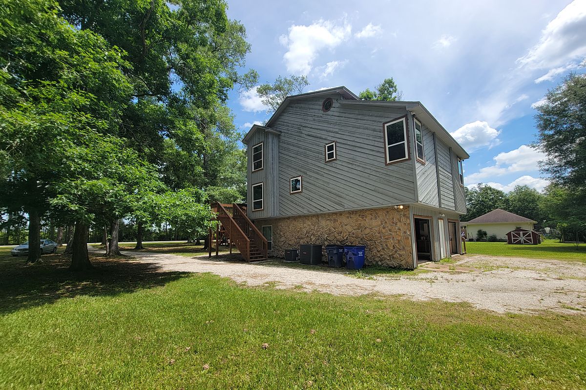 Two-story gray home with a stone lower level under tall shade trees in Cleveland, Texas